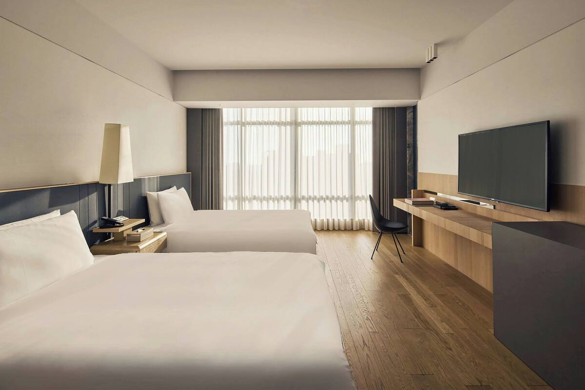 A hotel room with two beds, a desk, a television, and a large window with curtains.