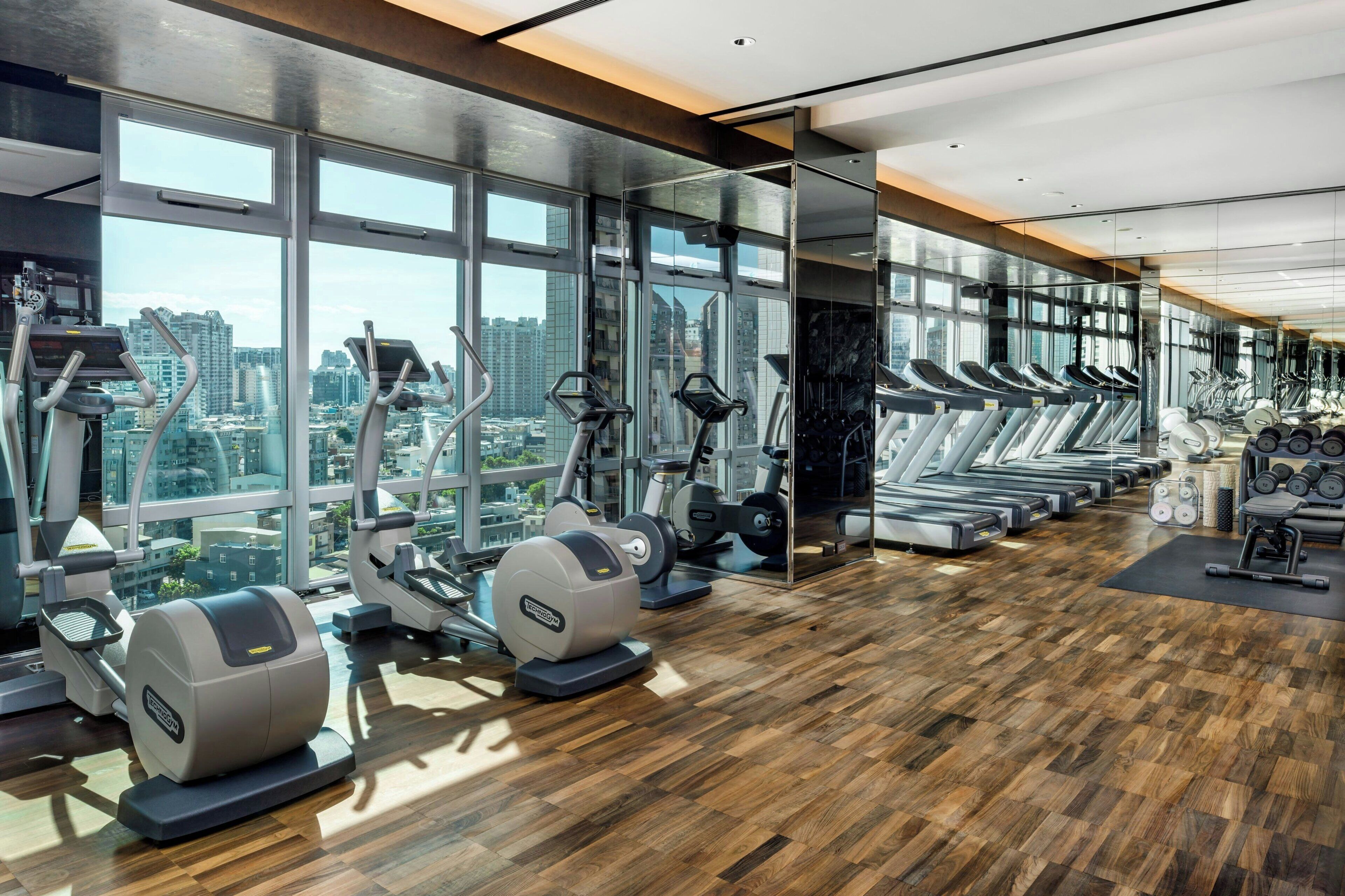 Een fitnessruimte met trainingsapparatuur, grote ramen en uitzicht over de stad.