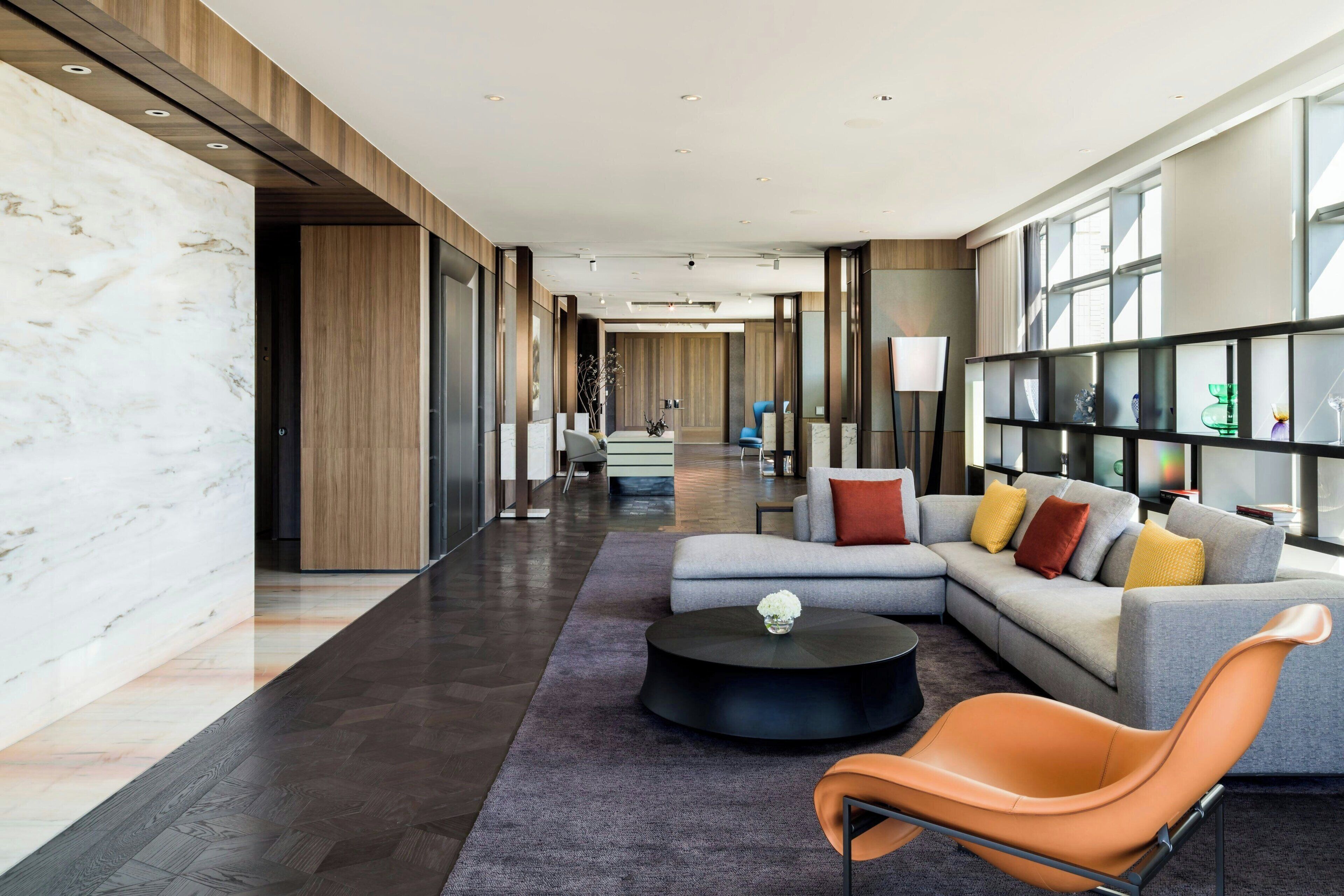 A modern hotel lobby with a marble wall, a sectional sofa with colorful cushions, a round black coffee table, and a unique orange chair.