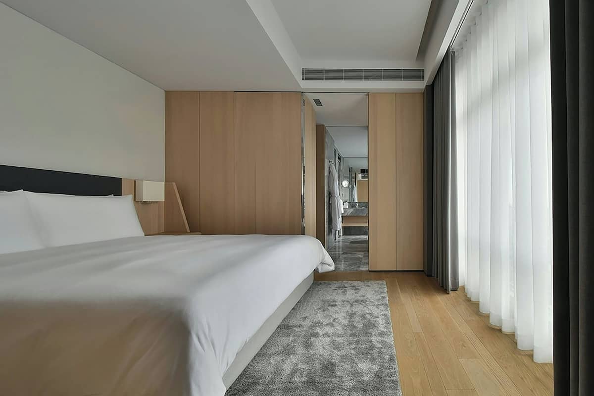 A modern hotel room with a large bed, wooden paneling, a walk-in closet, and a view of the hallway.