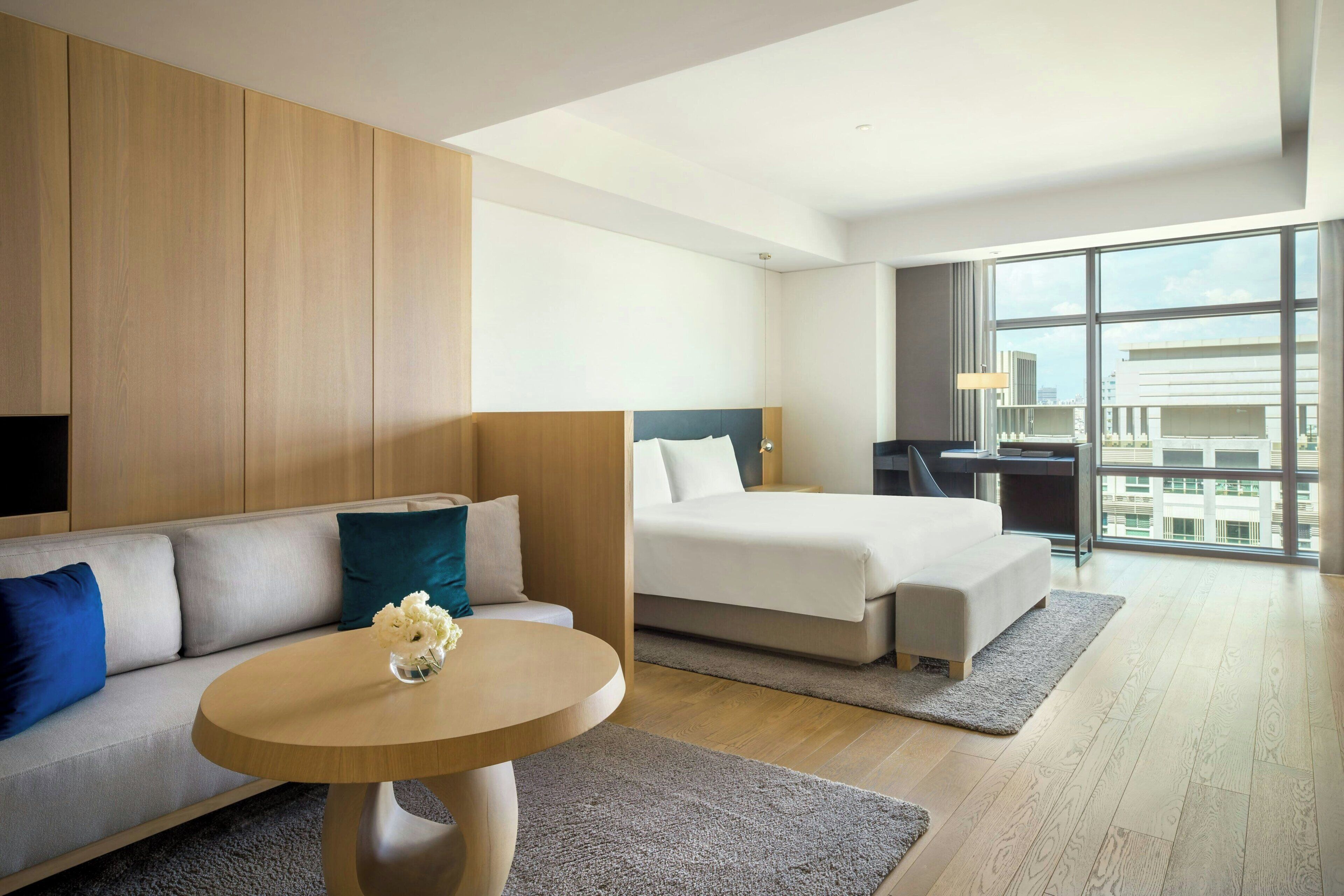 A modern hotel room with a sofa, a bed, a desk, and a large window offering a city view.