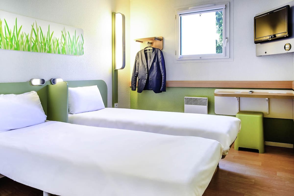 Quarto de hotel com duas camas, uma televisão, uma escrivaninha e uma janela.