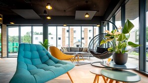 Un área de estar moderna con un sillón verde azulado, un sofá amarillo y un sillón colgante. Hay una planta en maceta sobre una mesa y vistas al exterior a través de amplias ventanas.