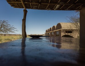 Un bâtiment rustique avec un toit de chaume et des fenêtres en arc de cercle, une table en bois avec un bol au premier plan, et un ciel bleu clair en arrière-plan.