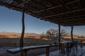 Un espace extérieur aménagé avec des tables et des chaises en bois, abrité par un toit de chaume, d’où l’on peut admirer un champ verdoyant et des montagnes lointaines.