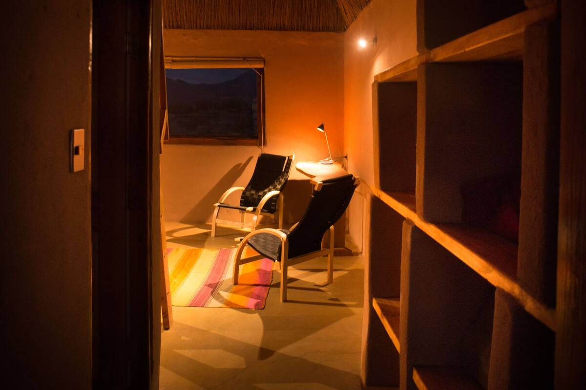 Ein schwach beleuchteter Raum mit gewölbter Decke, einem Schreibtisch mit Lampe, einem Stuhl und einem Fenster mit Blick auf eine Landschaft.