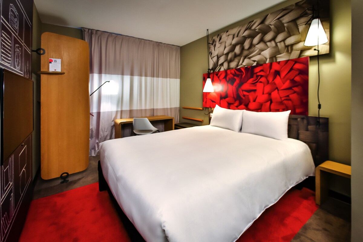 A hotel room with a large bed, a desk, a chair, a wardrobe, and a red carpet.