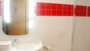 A bathroom with a white sink, mirror, and toilet, a shower area, and red-tiled walls. - ibis Mohammedia (Mohammedia)