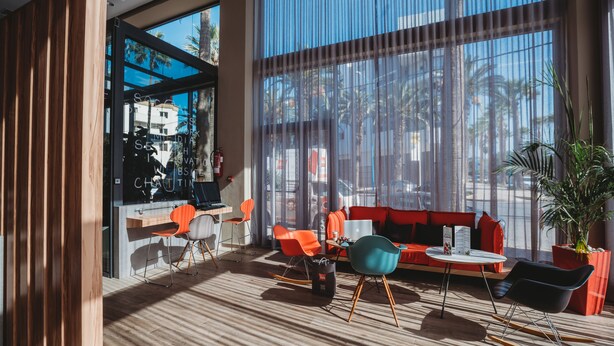 Un interior moderno con un separador de madera, una zona de estar con sillas anaranjadas y aguamarinas, un sofá rojo y un amplio ventanal con vista a palmeras.