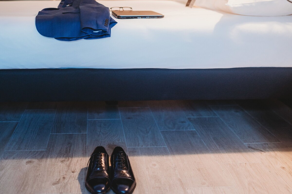 A hotel room with a neatly made bed, a pair of shoes on the floor, a blue bag, and a smartphone on the bed.