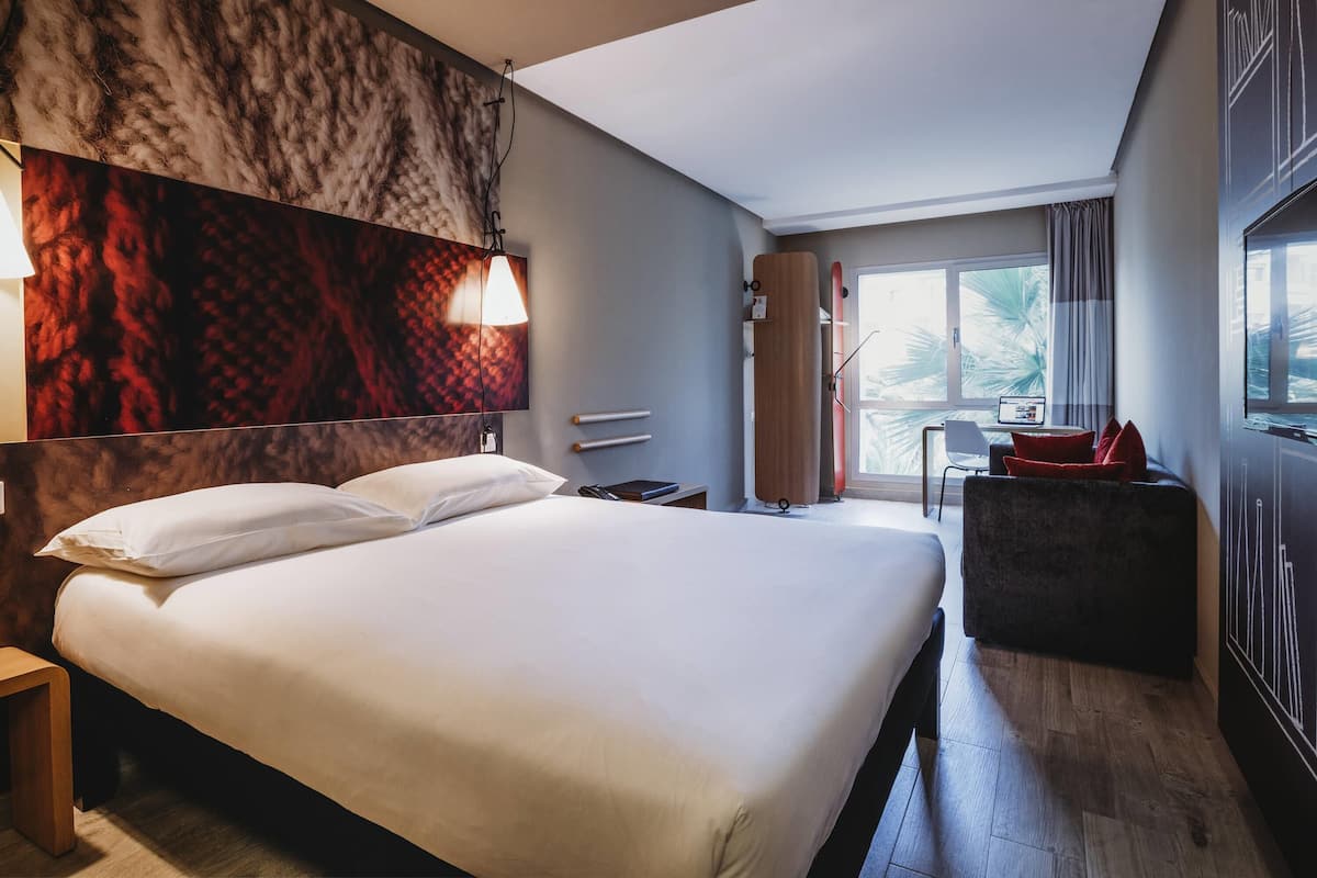 Habitación de hotel moderna con una cama grande, un escritorio con una computadora portátil, una silla y vistas a palmeras a través de la ventana.