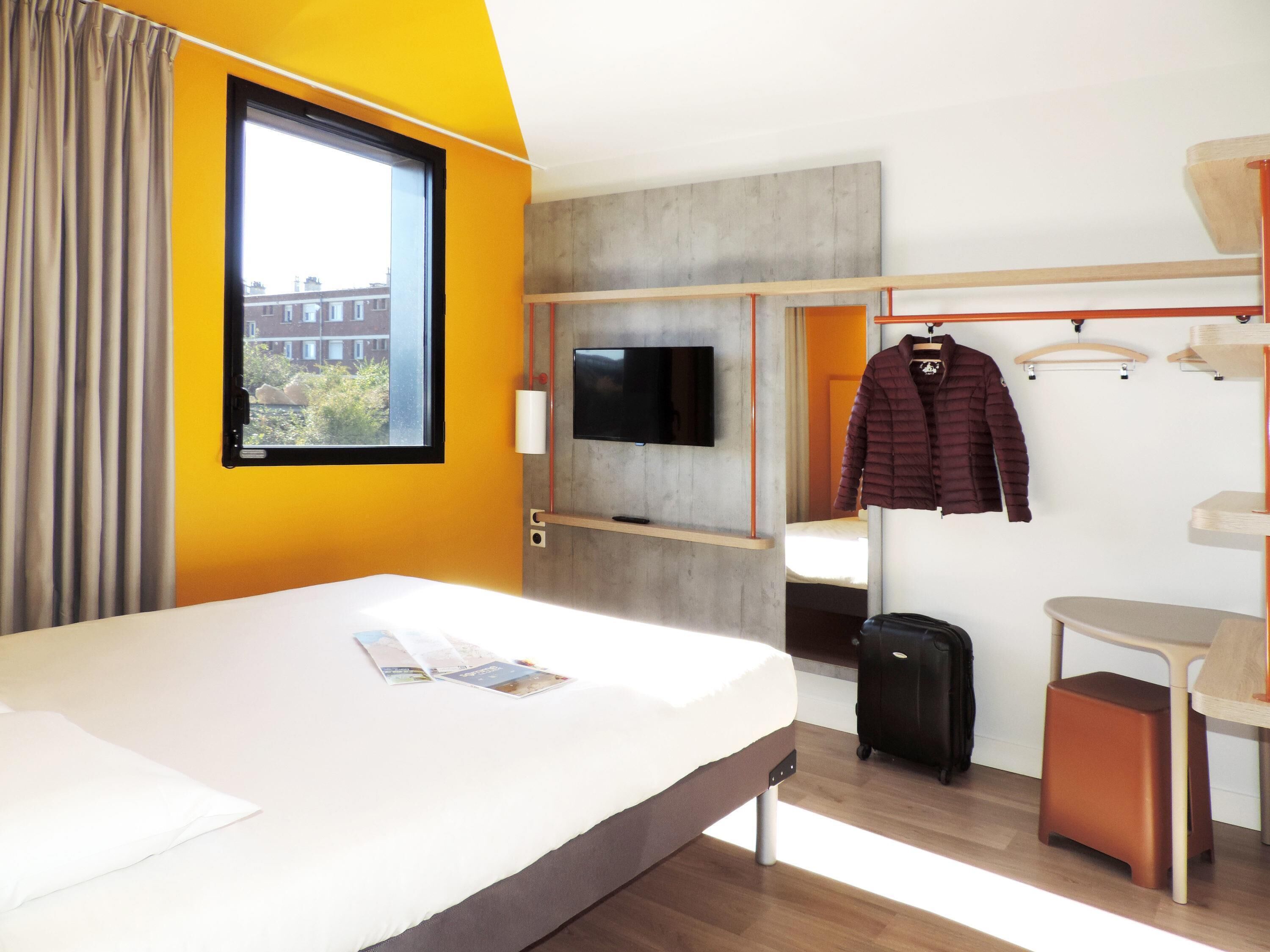 Une chambre d’hôtel avec un lit, une télévision, un porte-manteau, une valise et une fenêtre donnant sur des immeubles.