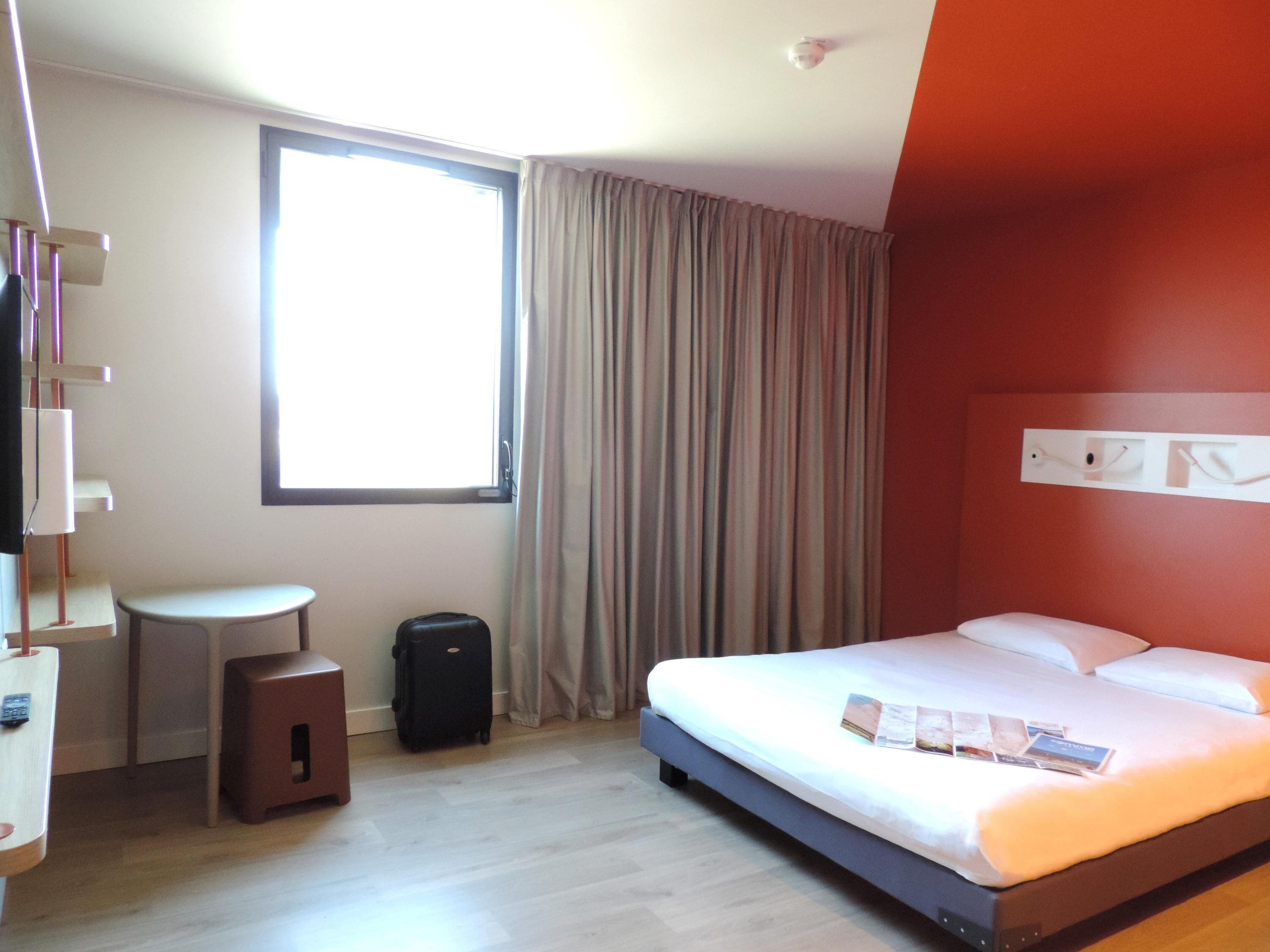 A hotel room with a bed, a small table, a suitcase, and a window with curtains.