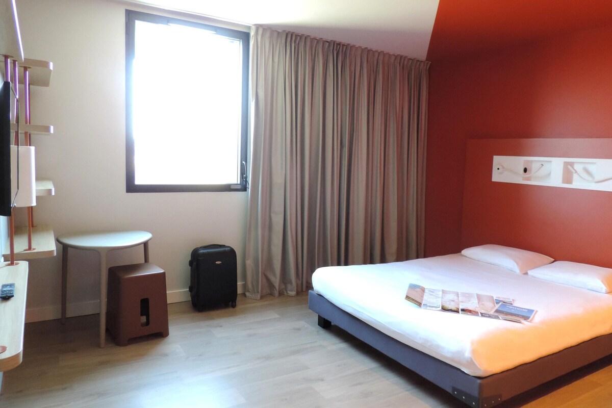 Une chambre d’hôtel avec un lit, une petite table, une valise et une fenêtre avec des rideaux.