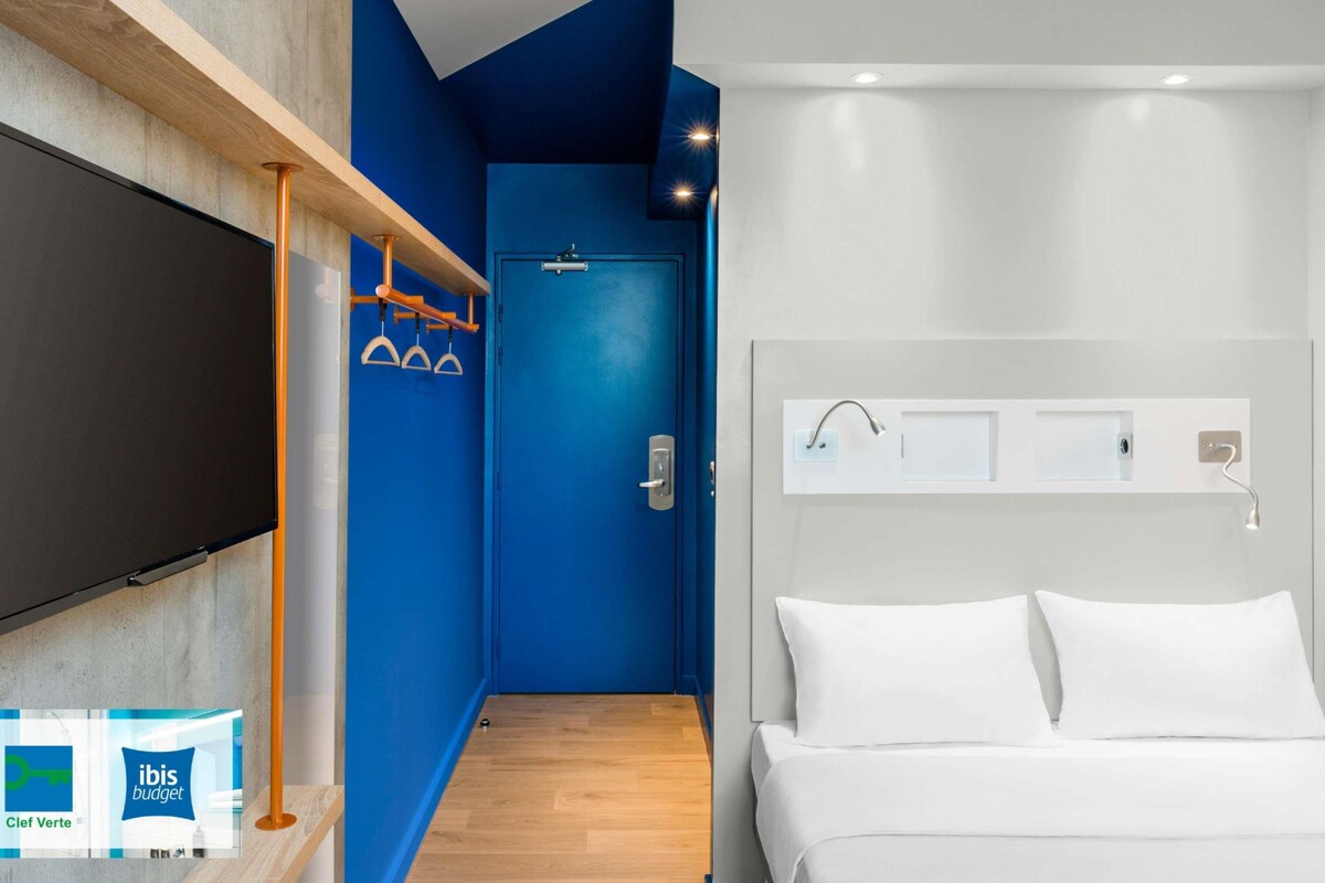 Une chambre d’hôtel avec un grand lit, un téléviseur fixé au mur, un espace douche et un mur d’accent bleu.