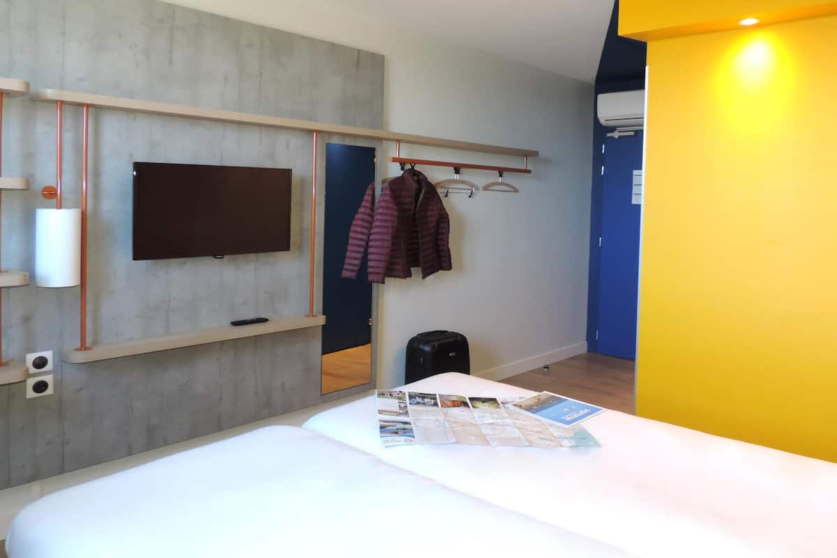 Une chambre d’hôtel avec un lit, un téléviseur à écran plat fixé au mur, un porte-manteau avec une veste et une valise posée sur le sol.