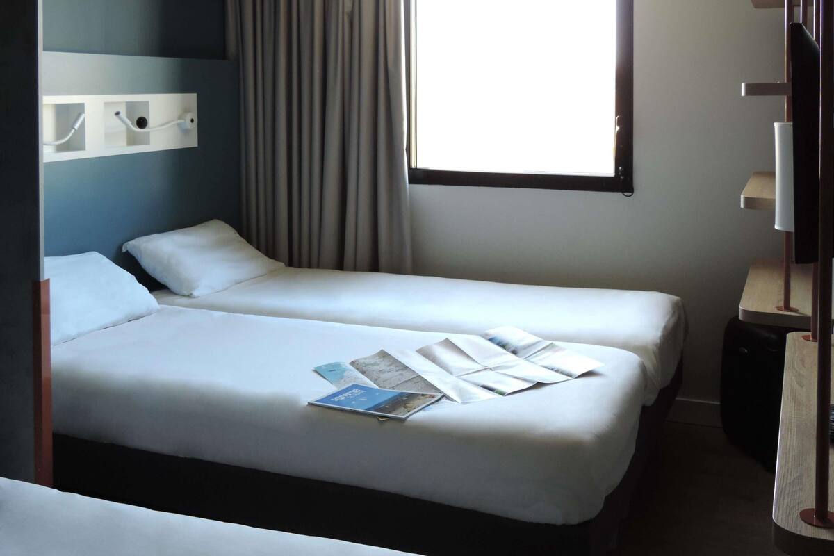 Une chambre d’hôtel avec deux lits, une fenêtre avec des rideaux, une table de chevet avec une lampe et une étagère fixée au mur.
