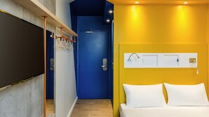 Quarto de hotel com uma cama, porta azul, parede branca com luminária e piso de madeira.