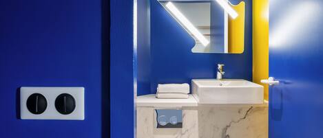 Un baño con pared azul, lavamanos blanco, espejo con luz en forma de piano y toallero.