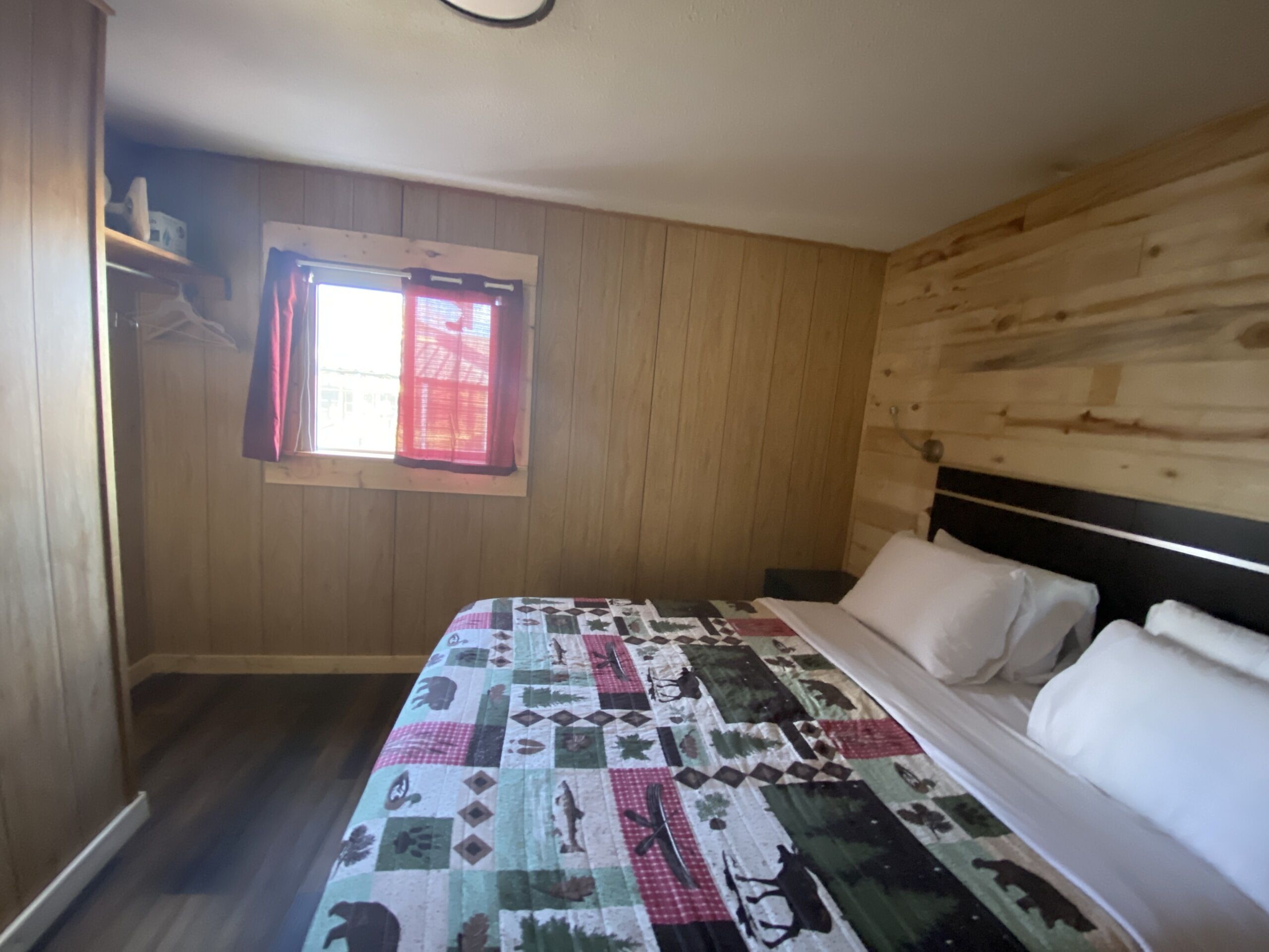 A bedroom with a wooden wall, a window with red curtains, a bed with a patterned bedspread, and a bedside table.