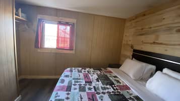 A bedroom with a wooden wall, a window with red curtains, a bed with a patterned bedspread, and a bedside table.