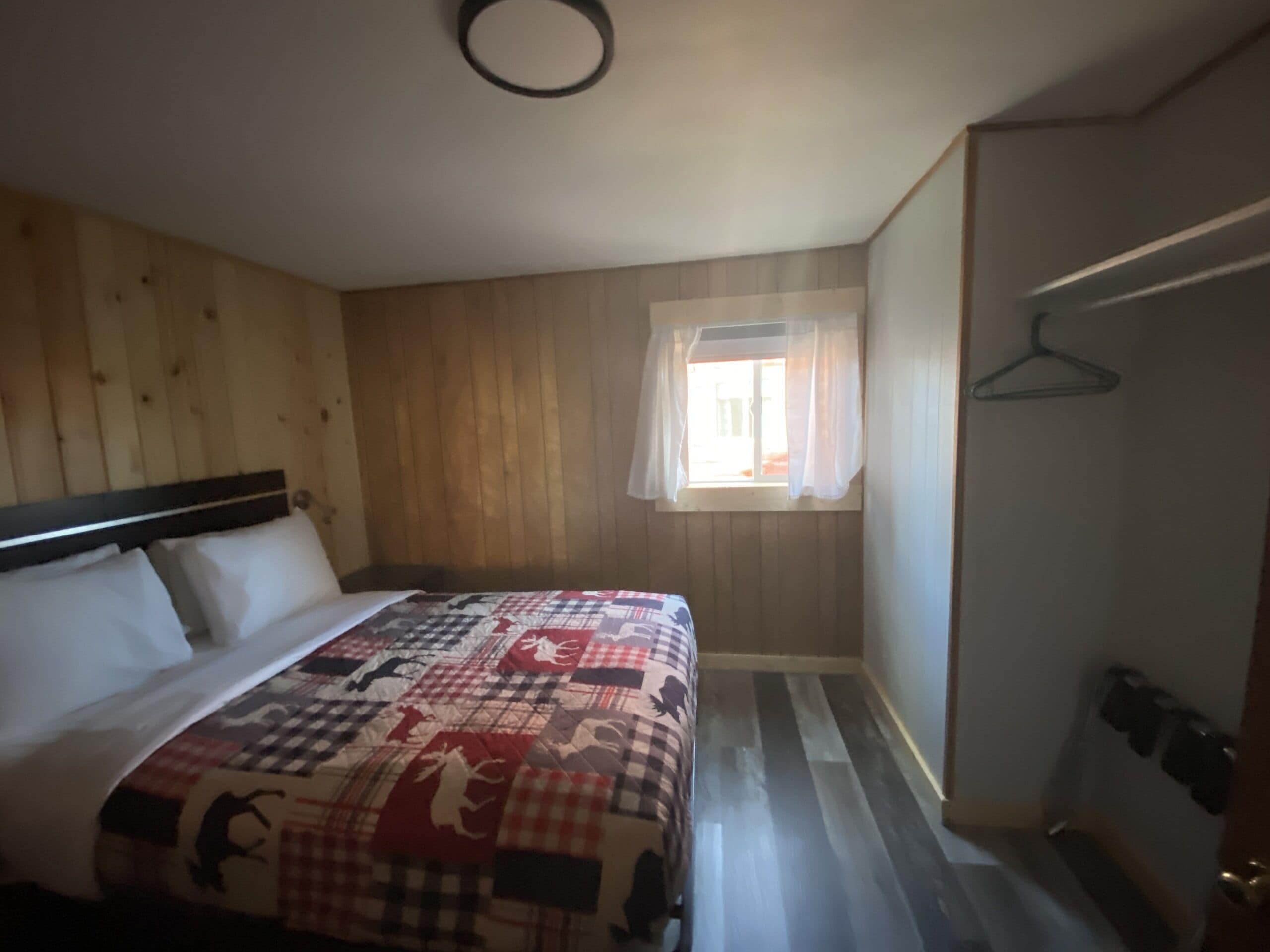 A bedroom with a wooden headboard, a quilted bedspread, a window with curtains, and a door with a coat hanger.