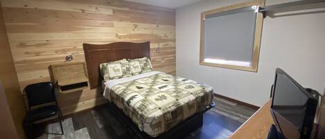A bedroom with a wooden headboard, a bed with a patterned bedspread, a bedside table, a desk with a computer monitor, and a window with blinds.