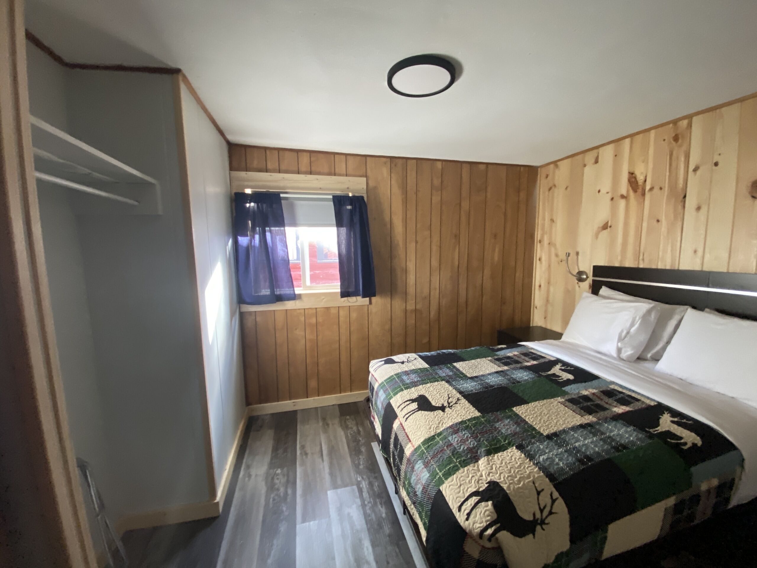 A bedroom with a wooden paneled wall, a window with curtains, a bed with a quilt, and a ceiling light.