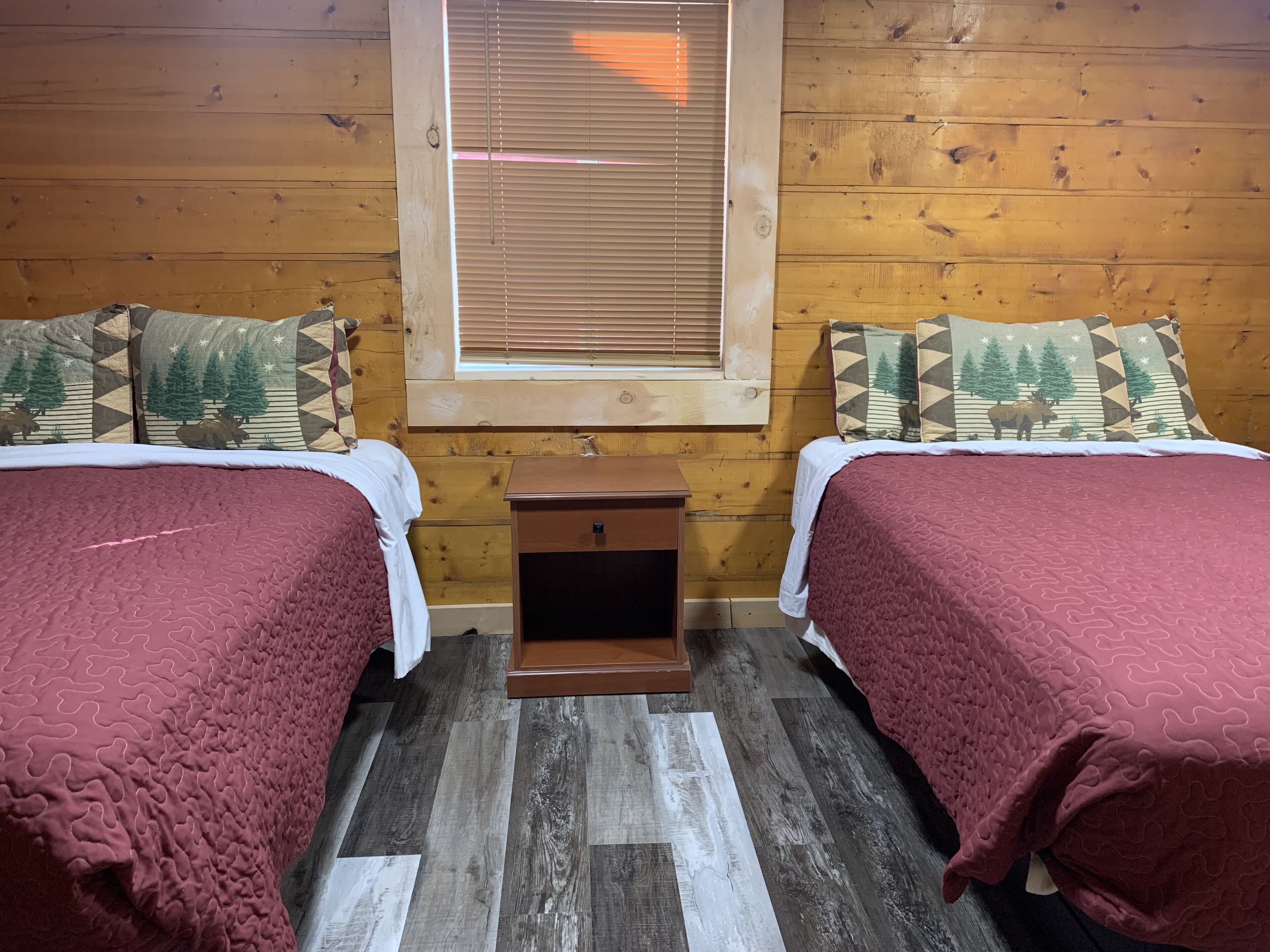 Une chambre avec deux lits, chacun recouvert d’une couette rouge, dotés de têtes de lit en bois et de coussins assortis. Une petite table de chevet en bois se trouve entre les deux lits, et une fenêtre est située au-dessus.