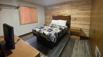Een slaapkamer met een houten hoofdbord, een bed met een quilt met patroon, een nachtkastje, een televisie op een houten bureau en een raam met jaloezieën.
