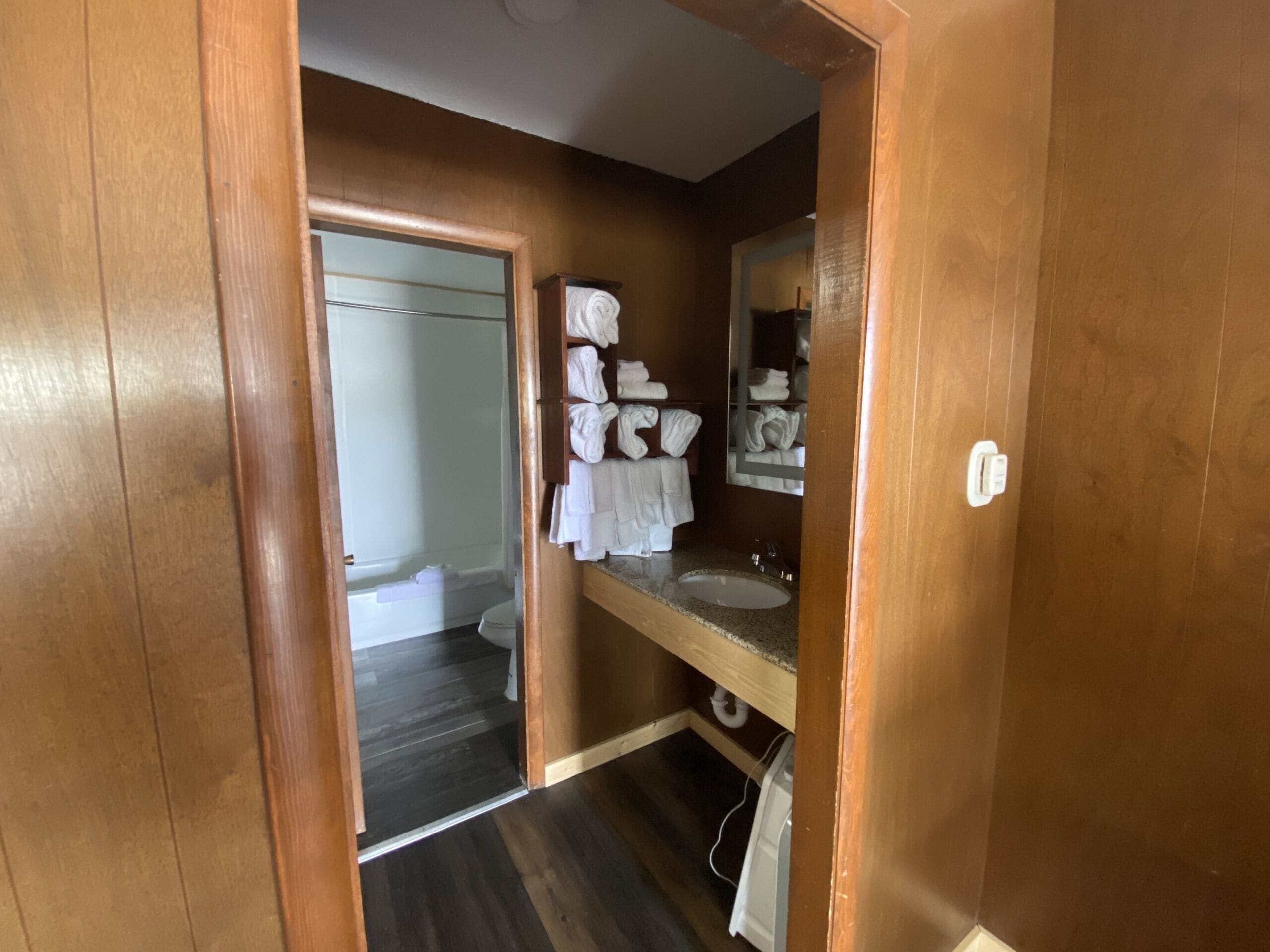 A bathroom with a wooden vanity, a sink, and a mirror. There is a towel rack with folded towels and a shower visible in the background.