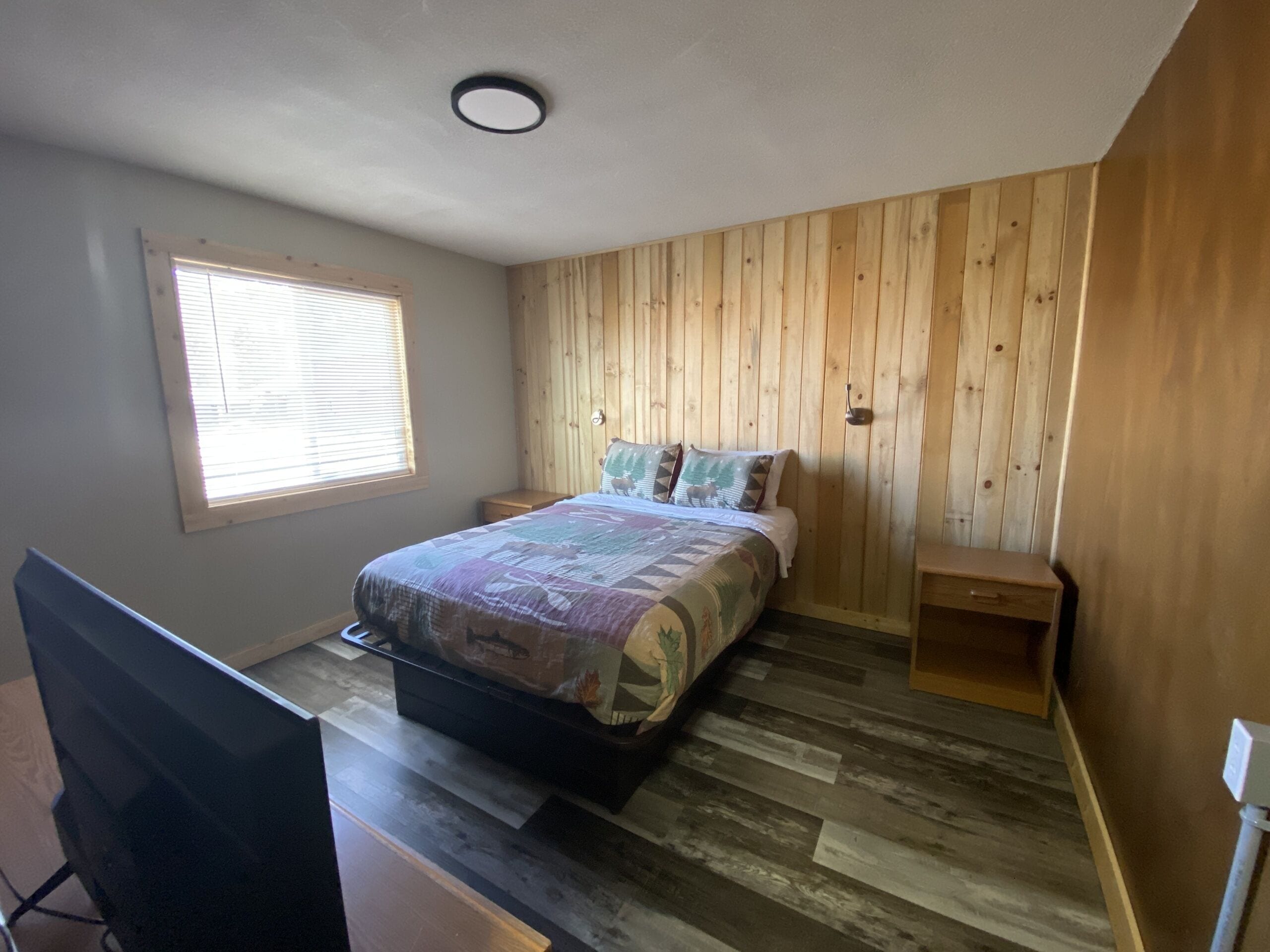 Un dormitorio con pared de madera, una cama con colcha estampada, una mesita de noche, un escritorio con computadora y una ventana con persianas.