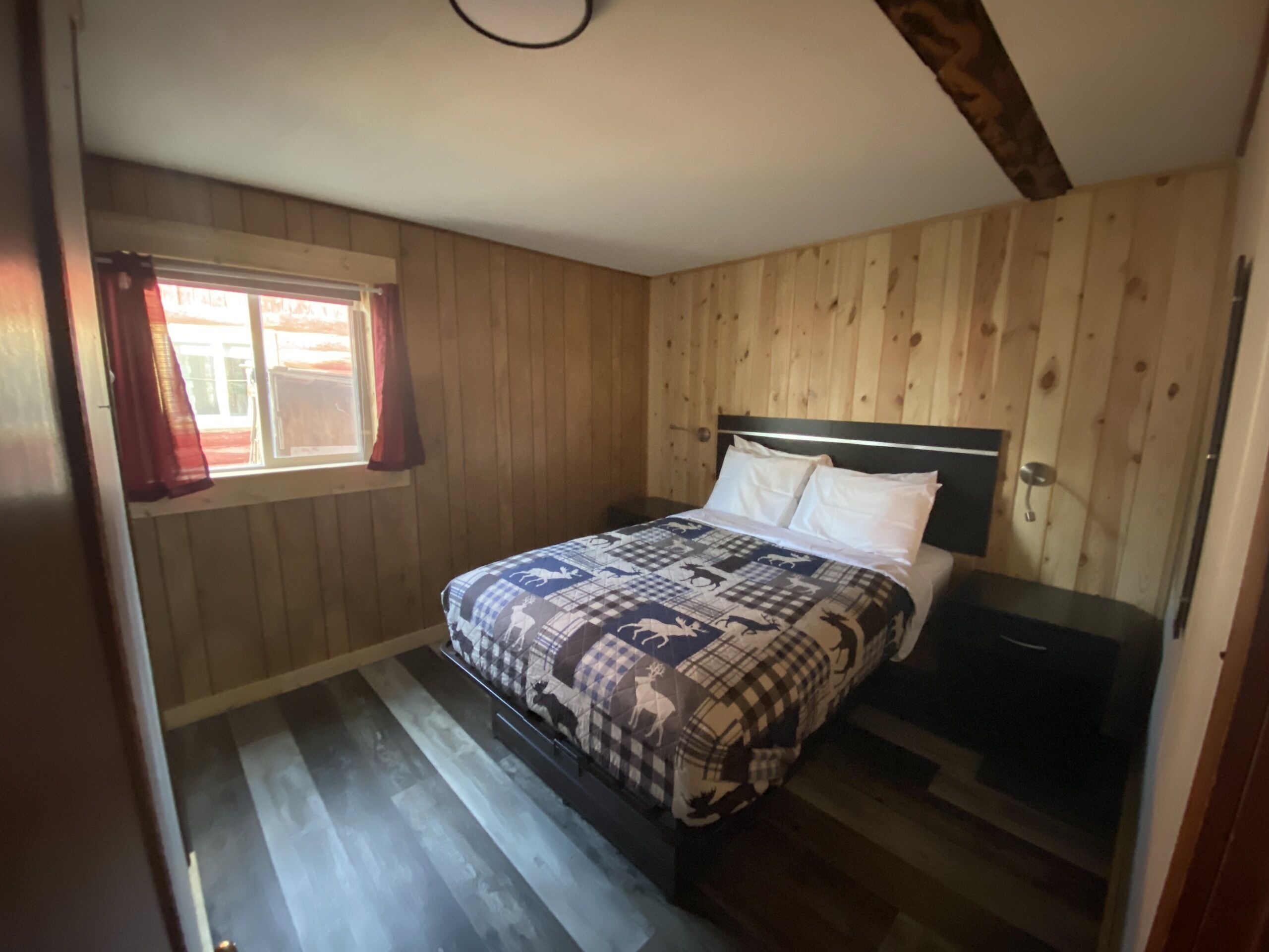A bedroom with a wooden paneled wall, a bed with a patterned bedspread, a nightstand, and a window with curtains.
