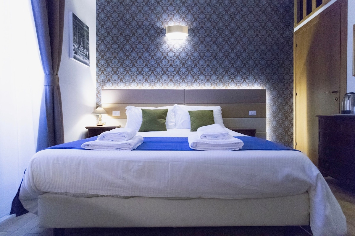 A neatly made bed with white and green pillows, a blue bedspread, and folded towels on the side.