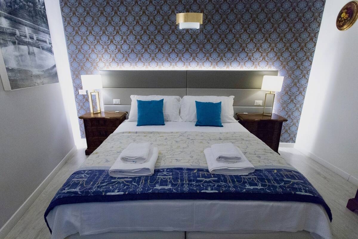 A hotel room with a large bed, two pillows, two bedside tables with lamps, and a patterned wallpaper.