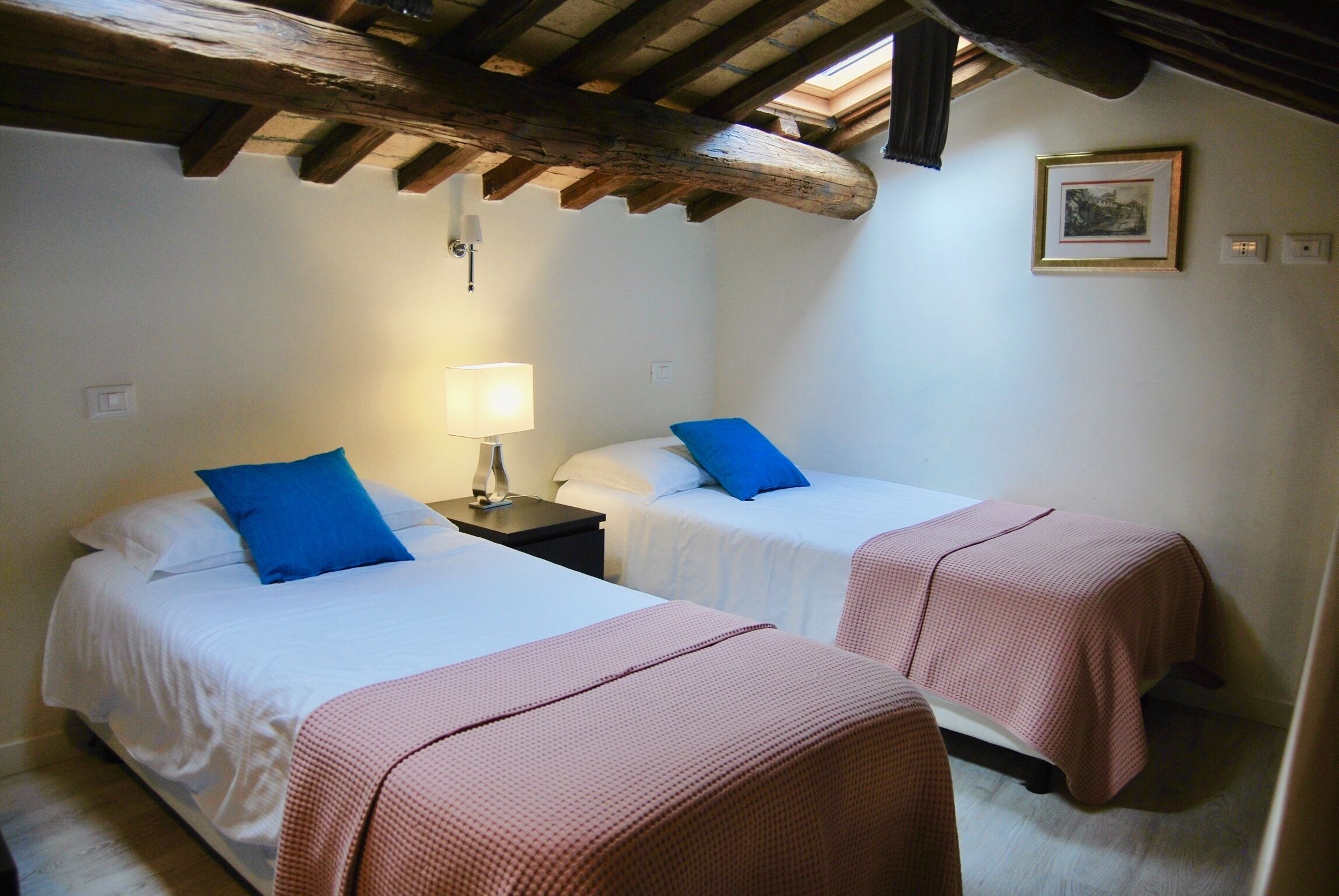 A room with two beds, each with a pillow and a blanket. There is a nightstand with a lamp between the beds. The room has wooden beams on the ceiling and a framed picture on the wall.