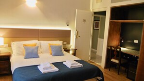 A hotel room with a large bed, two pillows, a blue bedspread, and a neatly made bed with towels. There is a wooden desk with a chair, a television, and a door leading to a bathroom. - Navona Residenza de Charme (Rome)