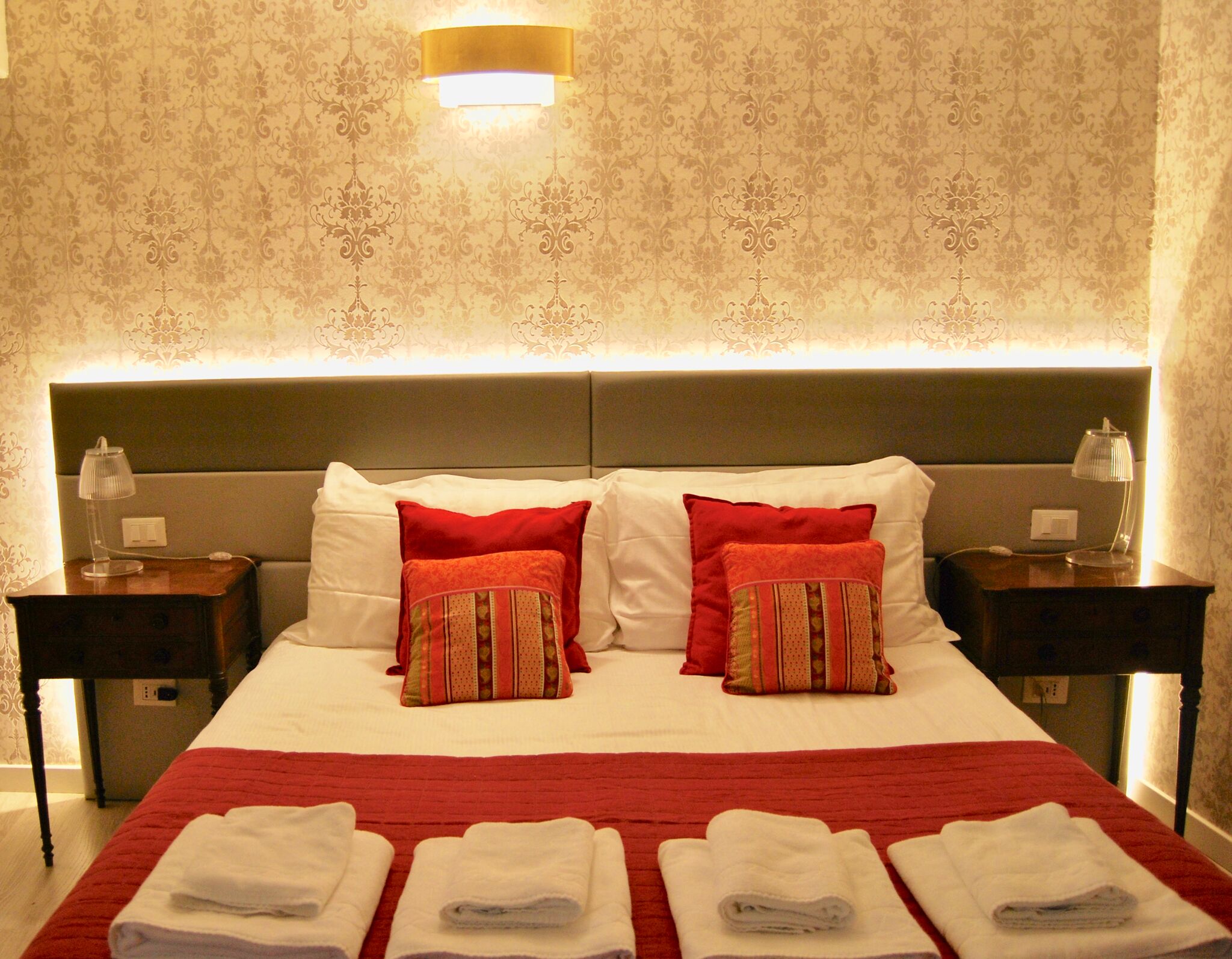 A neatly made bed with red and white bedding, two bedside lamps, and a patterned wallpaper.