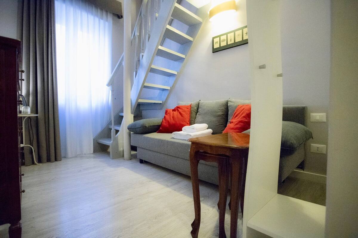 A modern living room with a grey sofa, red cushions, a wooden table, and a staircase.