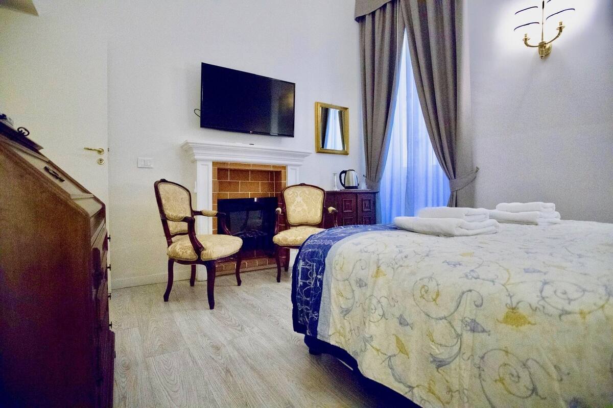 A hotel room with a bed, two armchairs, a television, a fireplace, and a window with curtains.