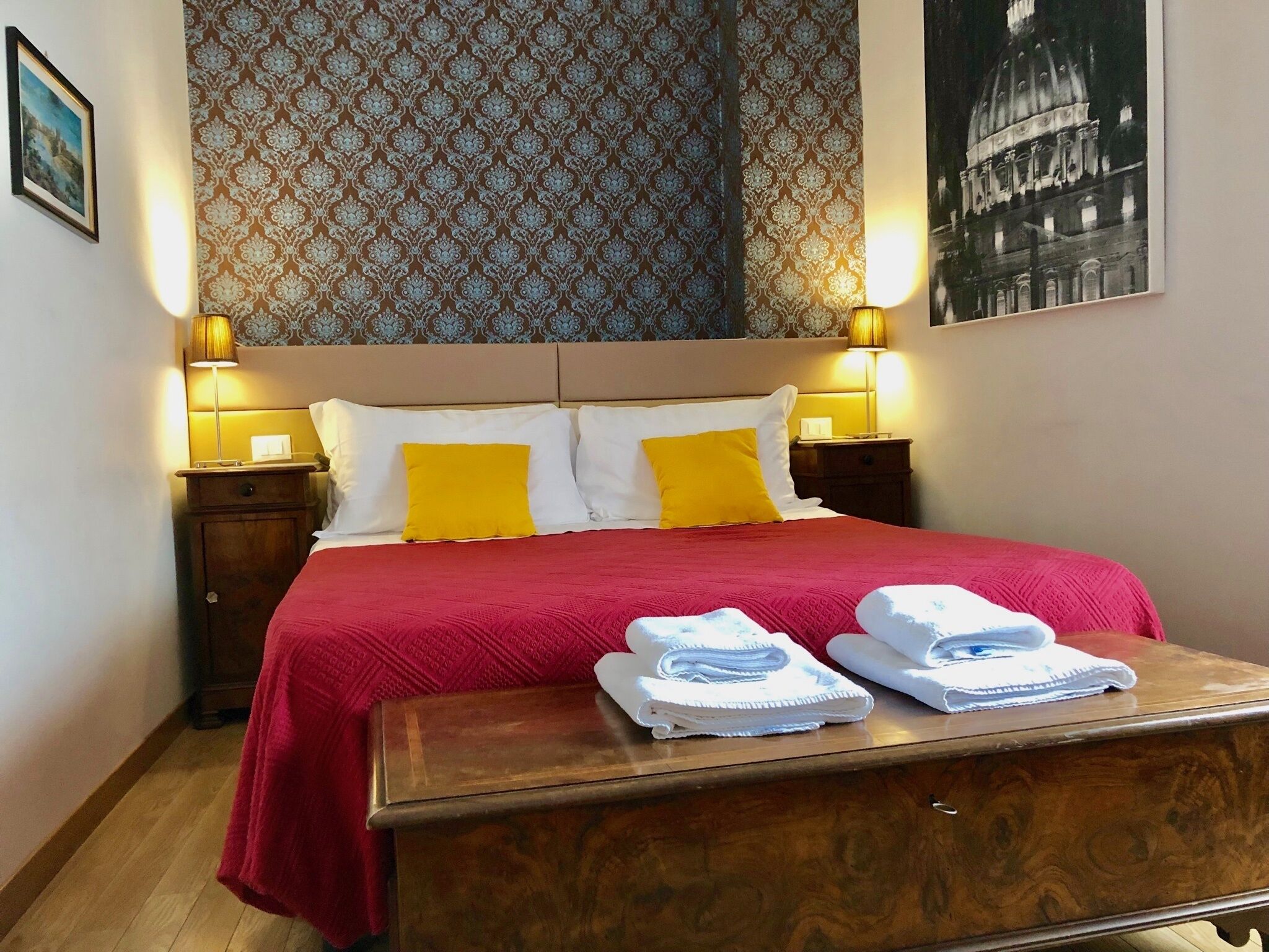 Habitación de hotel con cama, dos lámparas de noche, una mesita de noche de madera, una colcha roja, dos almohadas amarillas, un banco de madera con toallas, papel tapiz estampado y un cuadro en la pared.