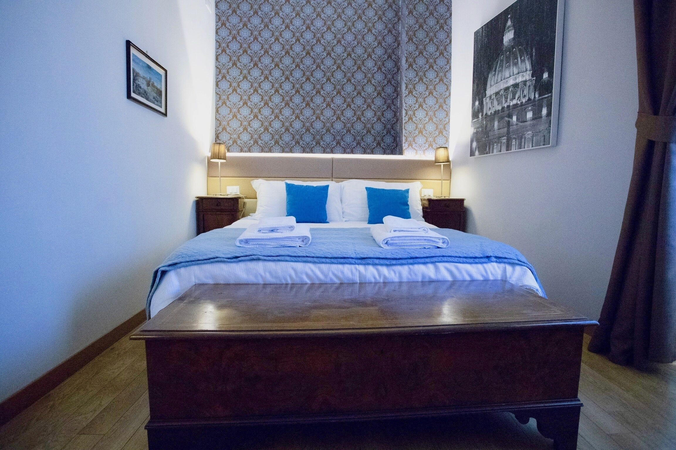 A neatly made bed with blue and white bedding, two bedside tables with lamps, patterned wallpaper, and a framed picture on the wall.