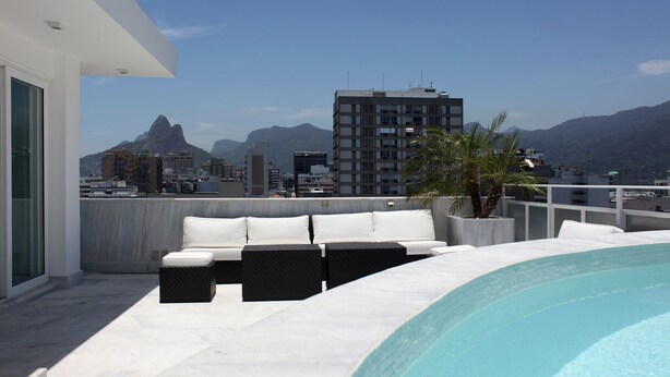 Area piscina sul tetto con una piscina a sfioro bianca, un divano da esterno bianco e nero e vista sullo skyline della città e sulle montagne sullo sfondo.