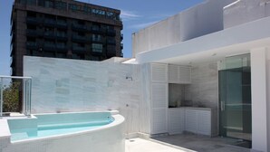 Area piscina esterna moderna con design curvo, cucina in vetro e vista su un grattacielo sullo sfondo.