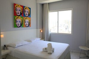 A hotel room with a large bed, a window with a city view, and a pop art painting on the wall.