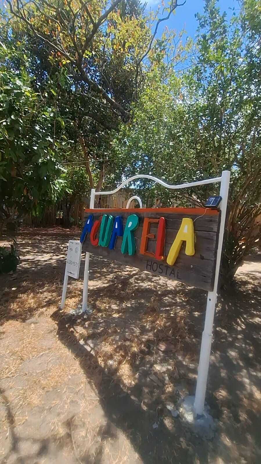A wooden sign with colorful letters spelling 'CUPREIA HOSTAL' in a natural setting with trees and dry grass.