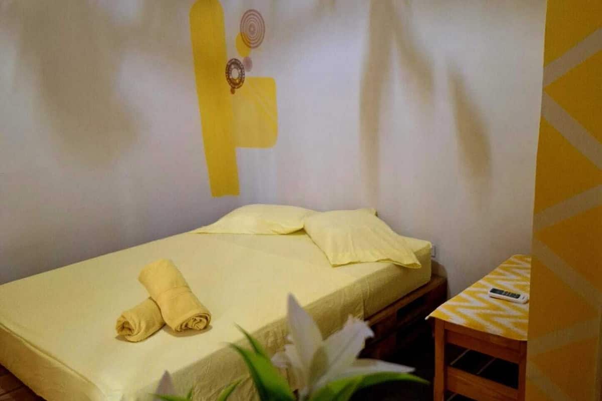 Una cama individual con colcha amarilla, una toalla enrollada y una mesita de madera con un control remoto.