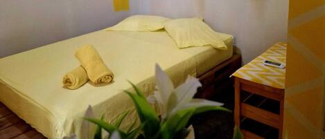 A single bed with a yellow bedspread, a rolled towel, and a small wooden table with a remote control.