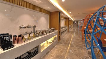 Espaço de buffet de hotel com bancada de mármore, estações de buffet e área de estar com cadeiras azuis.