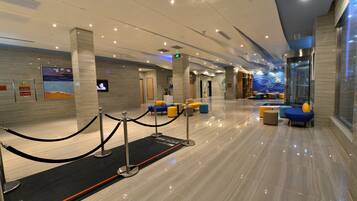 A modern hotel lobby with a reception desk, seating area, and a glass elevator.